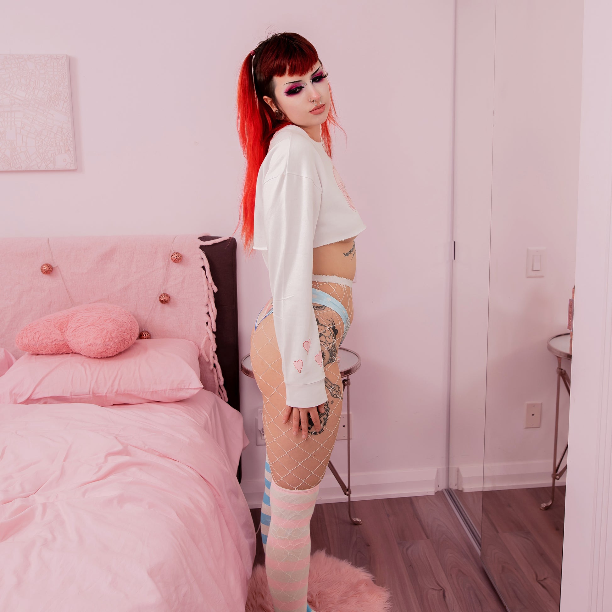 Person with red hair and tattoos standing in a bedroom with pink bed and walls.