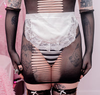 A close-up of a person wearing sheer black fishnet lingerie with horizontal cutout details, a white satin apron with lace trim, and black striped thong fabric, photographed against a soft pink background.